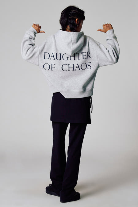 Grijze sweater met kap van Unwind, met de tekst "DAUGHTER OF CHAOS" op de rug, gedragen met een zwarte rok en broek.
