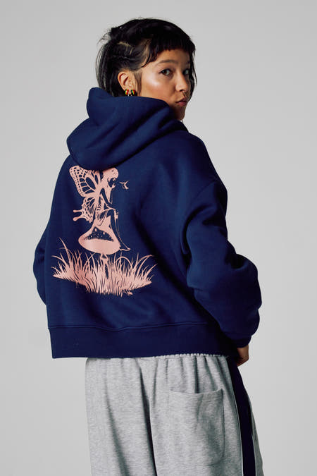 Blauwe sweater met kap van Unwind, met roze fee-print op de rug, gedragen over een grijze joggingbroek.
