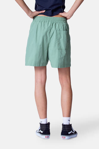 Groene short van Unwind, gezien van achteren, met elastische tailleband en achterzak, gecombineerd met witte sokken en zwarte Vans-sneakers.
