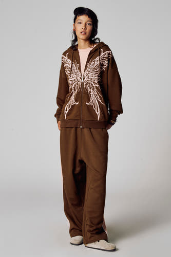 Bruine sweater van Unwind, met ritssluiting en roze vleugelmotief, gecombineerd met bijpassende joggingbroek en beige schoenen.
