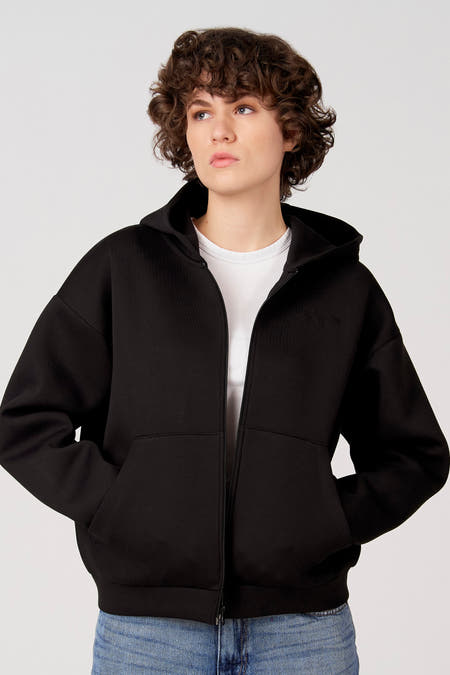 Zwart gilet van Unwind, met capuchon, lange mouwen en een ritssluiting, gedragen over een wit T-shirt en een blauwe jeans.
