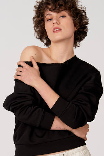 Zwarte sweater van Unwind, met een boothals die één schouder blootlaat, en gedragen met een crèmekleurige broek.
