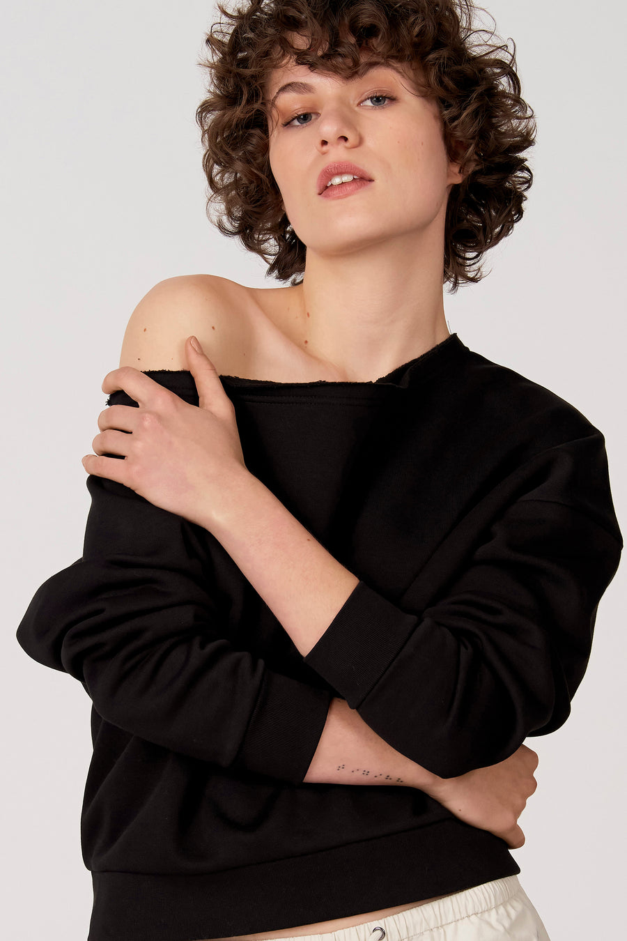 Zwarte sweater van Unwind, met een boothals die één schouder blootlaat, en gedragen met een crèmekleurige broek.
