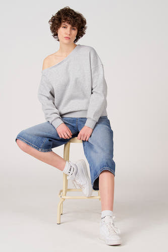 Grijze sweater van Unwind, met blote schouder gedragen, gecombineerd met wijde jeansbroek en witte sneakers op een kruk.
