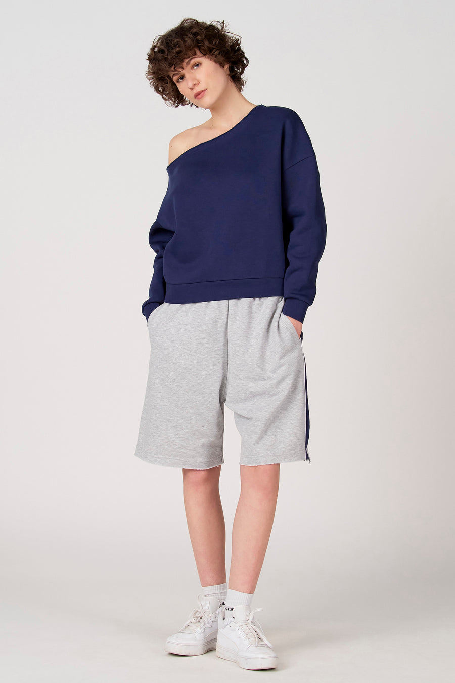 Blauwe sweater van Unwind, met blote schouder en gedragen met grijze shorts en witte sneakers.