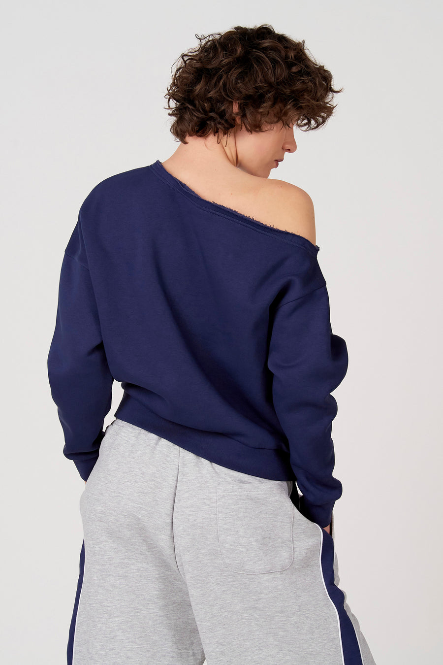 Blauwe sweater van Unwind, met afhangende schouder en gedragen met een grijze joggingbroek met blauwe streep.
