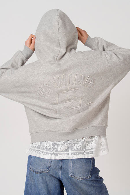 Grijze sweater met kap van Unwind, met sterren en de tekst "Unwind" op de rug, gecombineerd met een kanten bloes en jeans.
