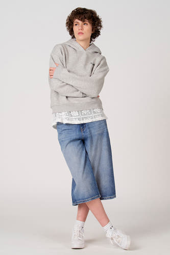 Sweat à capuche gris d'Unwind, porté les bras croisés sur un chemisier en dentelle blanche et une culotte en jean bleu.