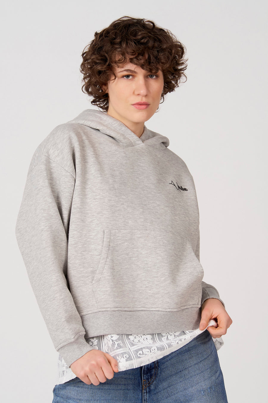 Grijze sweater met kap van Unwind, met trekkoord, een buidelzak en draagbaar over een witte kanten blouse en blauwe jeans.
