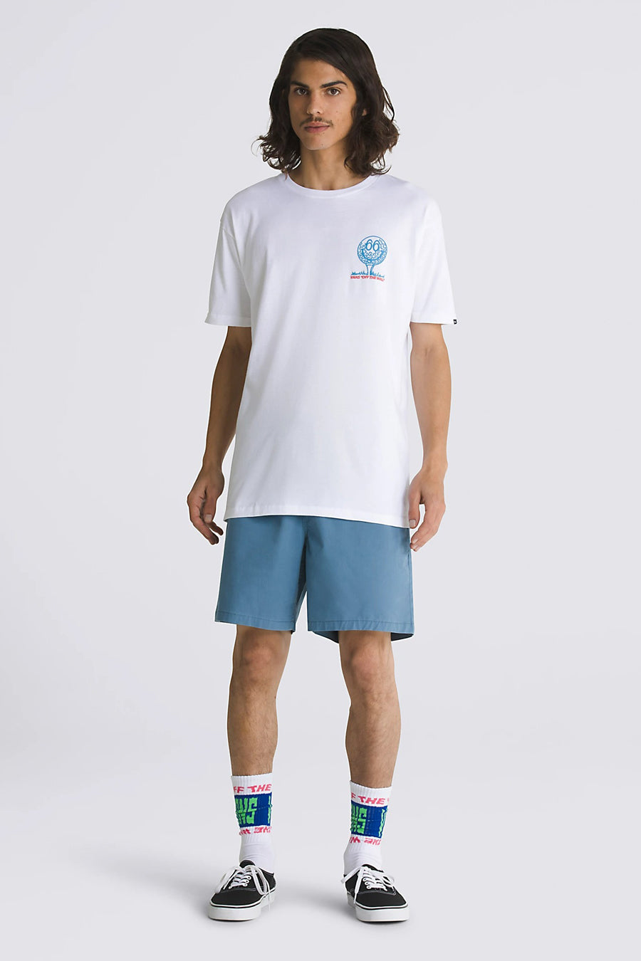 Een man staat gekleed in een wit T-shirt met print, blauwe shorts, witte sokken met blauwe en groene letters, en zwarte sneakers.