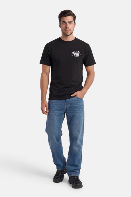 Zwart T-shirt van VANS met een wit ovaal logo op de borst, gedragen door een man met blauwe jeans en zwarte sneakers.