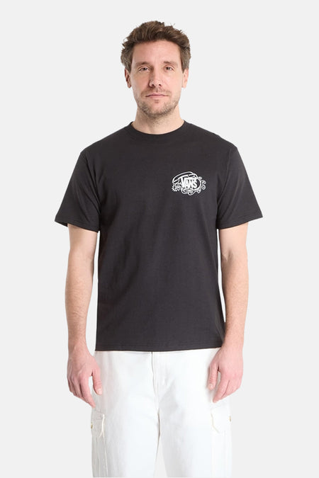 Zwart T-shirt van VANS met wit, gestileerd logo op de linkerborst, gedragen door een man in een witte broek.