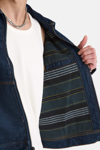 Donkerblauwe jeansjas van VANS “OFF THE WALL”, met een geruite voering en gedragen over een wit hemd met zilveren ketting.
