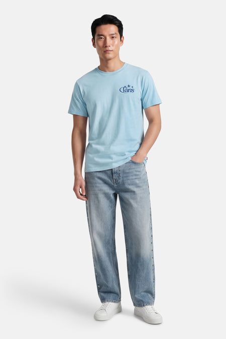 T-shirt bleu clair de VANS avec logo Vans bleu foncé et étoiles sur la poitrine, porté par un homme avec un jean clair et des baskets blanches.