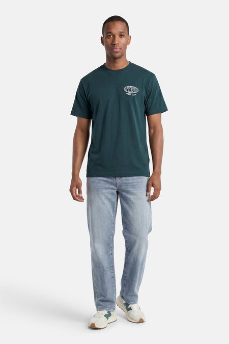 Groen T-shirt van VANS met een wit ovaal logo en 'FOREVER ON TOUR SINCE 1966' tekst, gedragen door een man met een lichte spijkerbroek en beige sneakers.