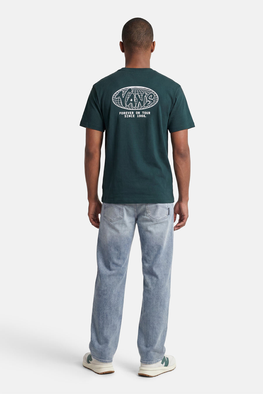 Groen T-shirt van VANS "OFF THE WALL" met wit "VANS" logo in wereldbolraster en tourtekst op de rug, gedragen met een lichte spijkerbroek en sneakers.