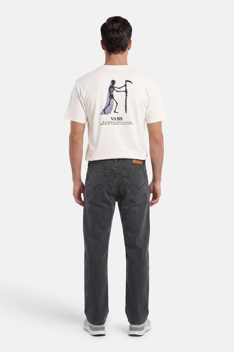 Ecru T-shirt van VANS “OFF THE WALL” met skeletprint en tekst op de rug, gedragen door een man in geribbelde jeans en sneakers.