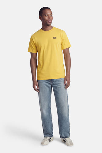 Geel T-shirt van VANS “OFF THE WALL”, gedragen met jeans en crèmekleurige sneakers met zwarte strepen.