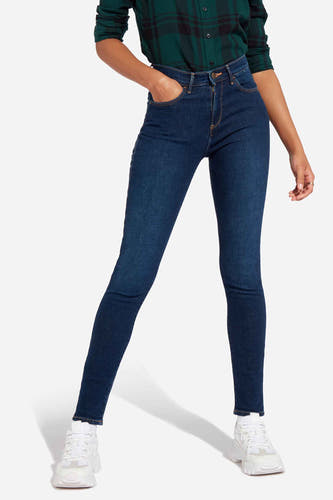 Jean skinny en denim de Wrangler®, en délavage bleu foncé, combiné avec une chemise à carreaux verte et des baskets blanches.