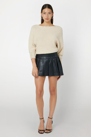 Jupe courte noire de Guess, avec des plis et des détails décoratifs en cuir, associée à une chemise beige et des baskets noires.