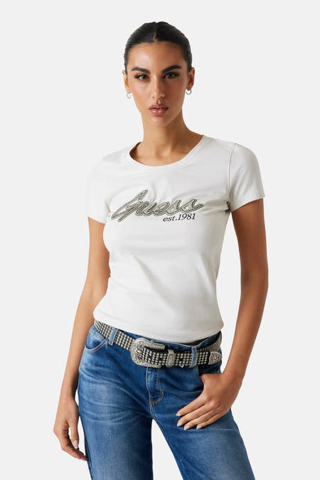 Wit T-shirt met korte mouwen van Guess, met glitters en de tekst "est. 1981", gedragen met een jeans en riem.
