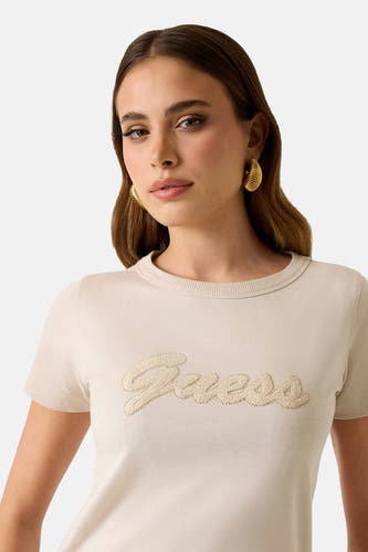 T-shirt beige à manches courtes de Guess, avec logo brodé sur le devant, porté avec des boucles d'oreilles dorées.