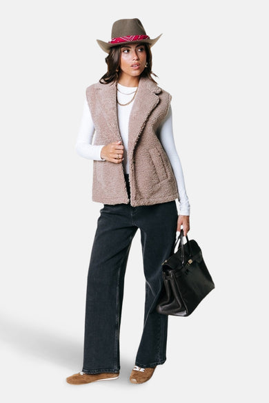 Beige bodywarmer van COLOURFUL REBEL, gedragen over een wit shirt met lange mouwen, gecombineerd met een zwarte broek en bruine schoenen, met een zwarte tas.