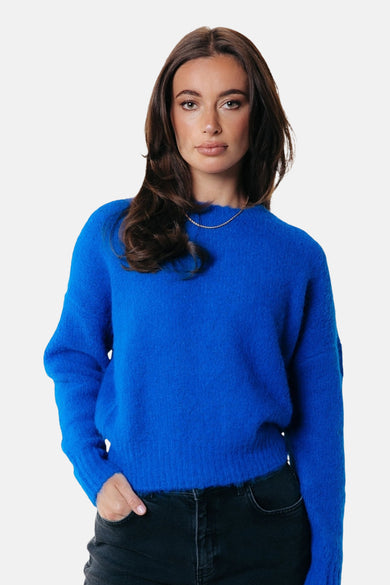 Blauwe trui met ronde hals van COLOURFUL REBEL, gedragen met een zilveren ketting en een zwarte broek.
