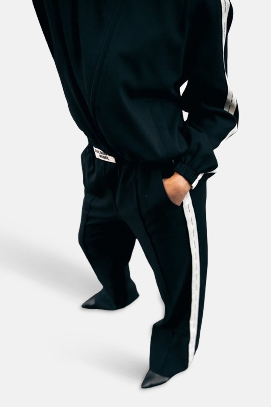 Pantalon noir de COLOURFUL REBEL, avec une bande blanche sur le côté et la main dans la poche.