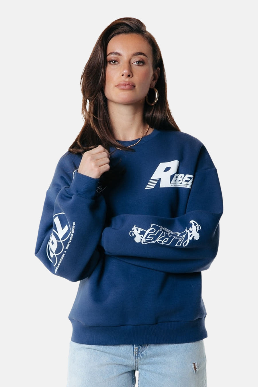 Pull-over bleu de COLOURFUL REBEL, avec des impressions de logo blancs sur les manches et la poitrine, porté avec des boucles d'oreilles dorées.