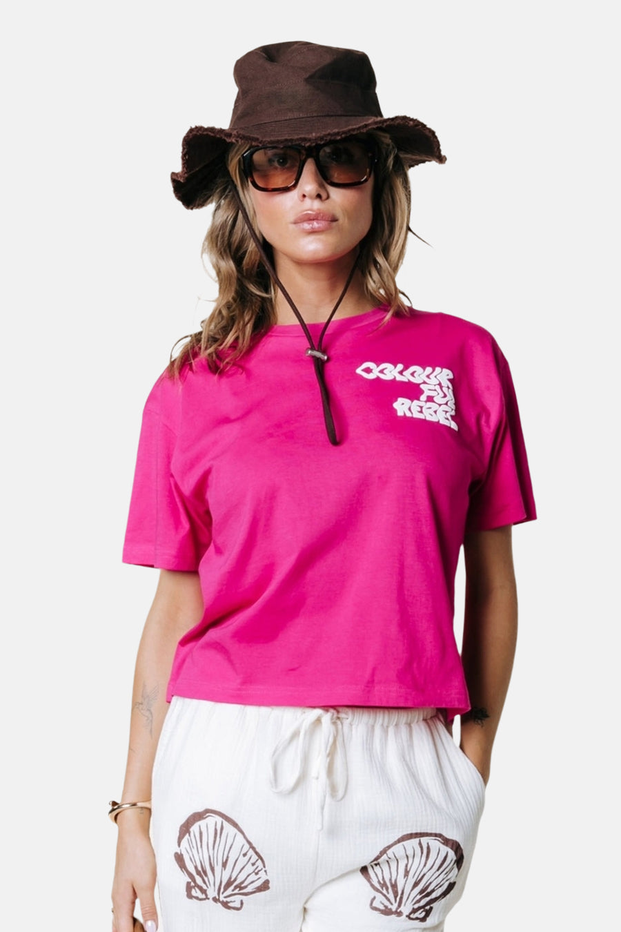 Roze T-shirt van COLOURFUL REBEL met witte logoprint, gedragen door model met hoed en korte broek met schelpenprint.