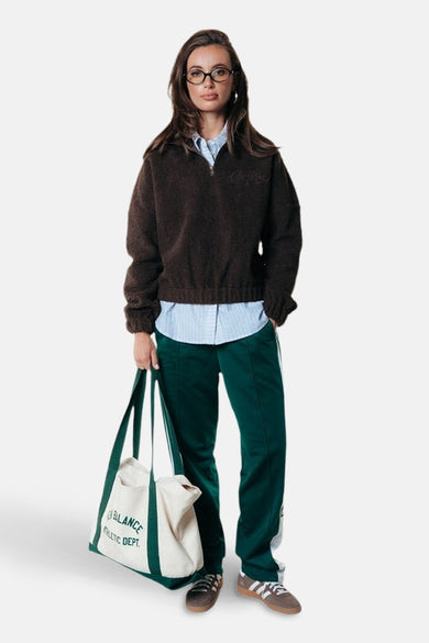Bruine fleece van COLOURFUL REBEL, gedragen met een gestreept overhemd, groene joggingbroek met witte strepen en een canvas tas.