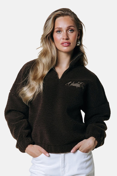 Bruine fleece van COLOURFUL REBEL, met een korte ritssluiting en geborduurd logo, gecombineerd met een witte broek en gouden oorbellen.
