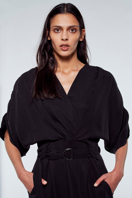 Robe longue noire de YŪGEN avec col V, détail ce ce et manches larges, les mains dans les poches.