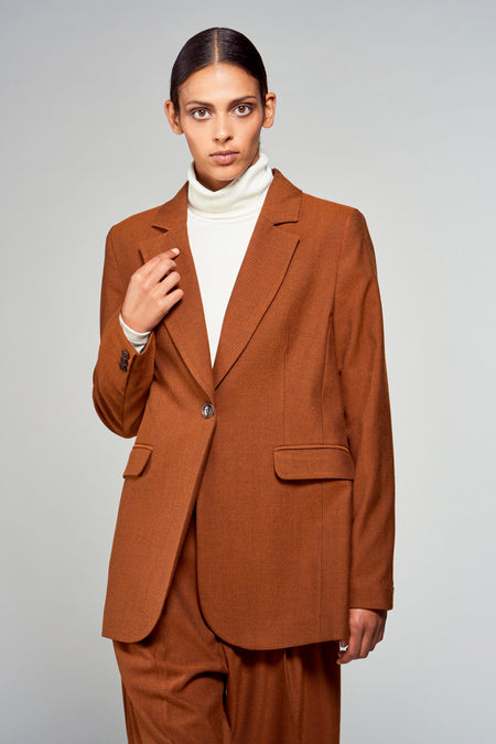 Bruine blazer van YŪGEN, gedragen over een witte coltrui, met een enkele knoop en paspelzakken.