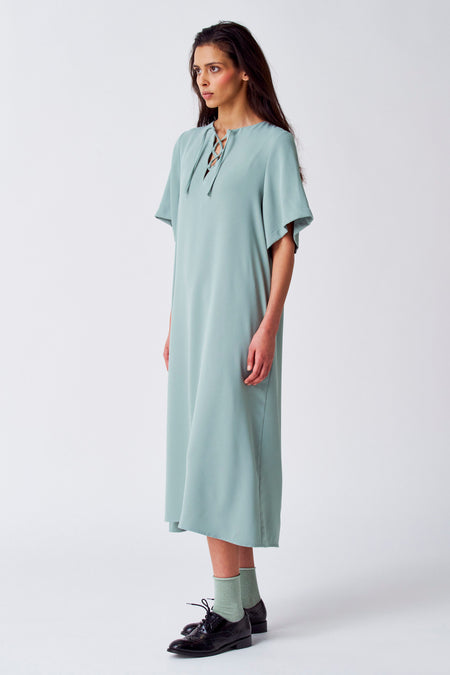 Groene jurk met korte mouwen van YŪGEN, met vetersluiting en gedragen met groene sokken en zwarte schoenen.
