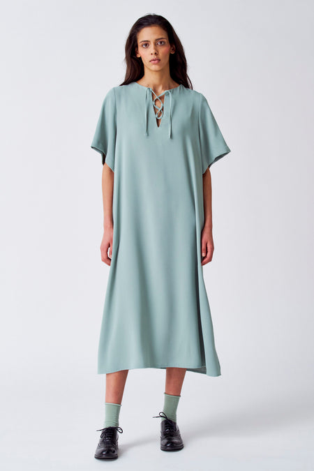 Groene jurk met korte mouwen van YŪGEN, met veterdetail bij de hals en zichtbare groene sokken met zwarte schoenen.
