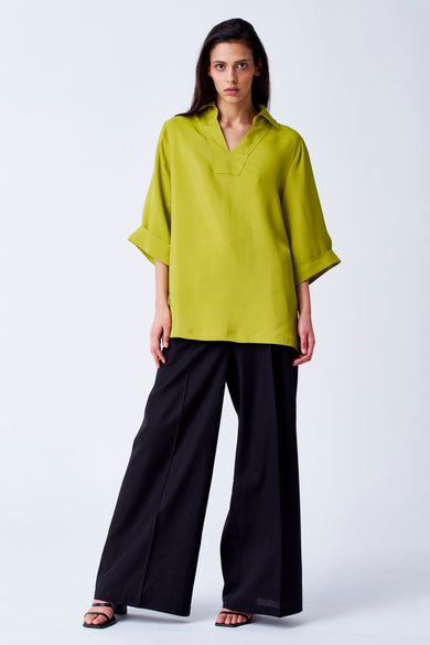 Gele blouse met driekwart mouwen van YŪGEN, gedragen met een zwarte, wijd uitlopende broek en sandalen.
