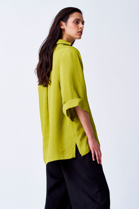 Gele blouse van YŪGEN met driekwart mouwen, gecombineerd met een zwarte broek en een effen achtergrond.

