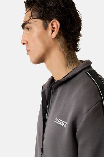 Grijs vest van Guess met rits en witte merklogo, detail op de schouder en nek tatoeage.
